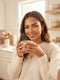 A candid morning selfie of a woman with perfectly defined, symmetrical eyebrows holding a coffee mug in a sunlit kitchen.